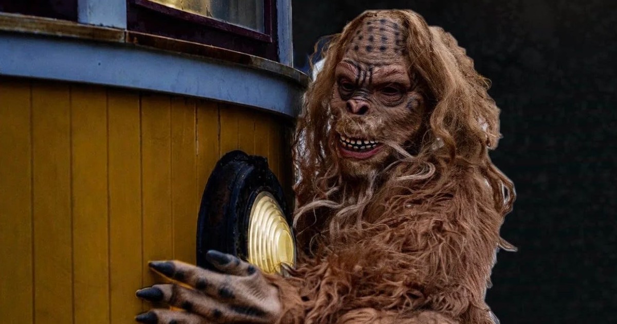 A Sasquatch in a shaggy costume leans against a yellow trolley beside its glowing headlight