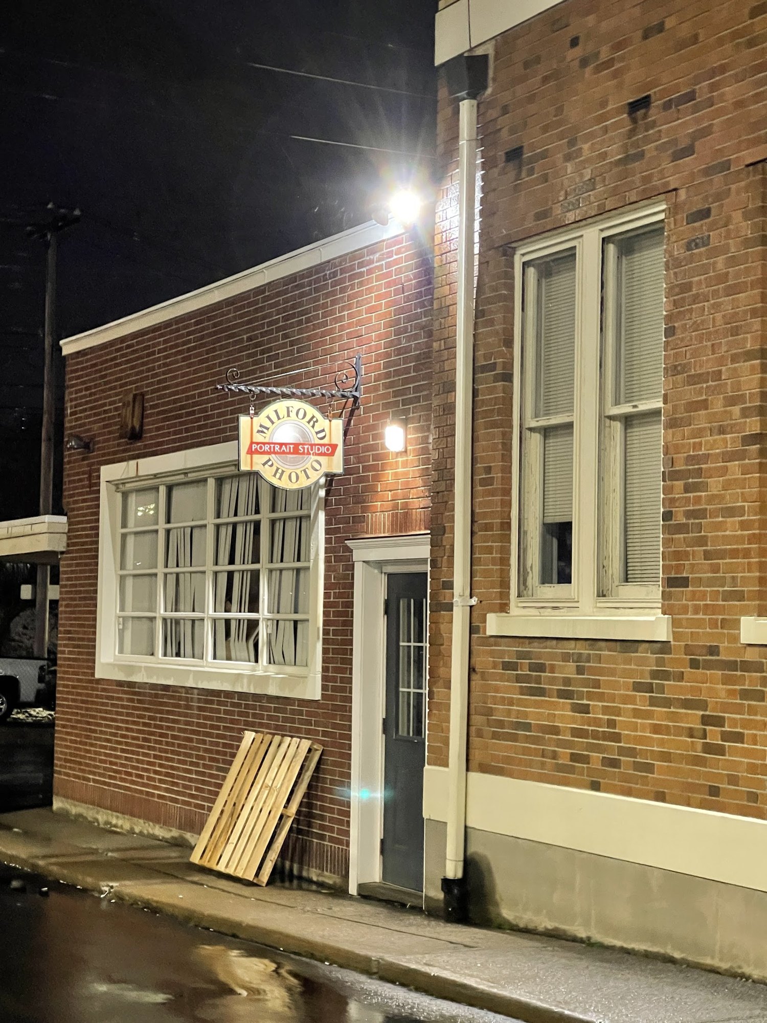 The brick side entrance of Milford Photo on River Street at night, with its hanging Milford Portrait Studio sign lit up
